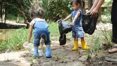 一个亚洲小孩，带着一家人在一条公共河流的边缘用黑色袋子捡起垃圾和塑料瓶。